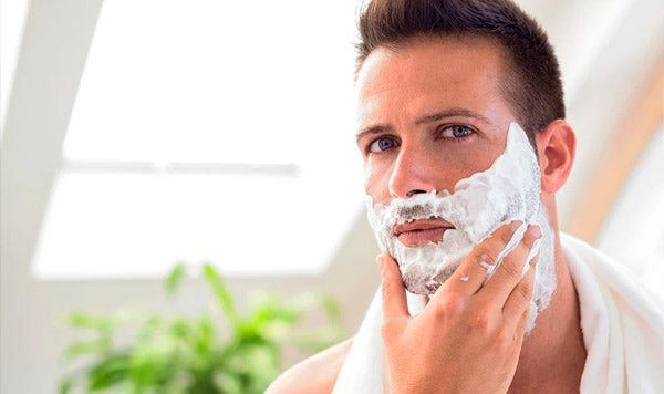 A man with white shaving foam on his face applying a shaving towel, shown in a bright bathroom setting.