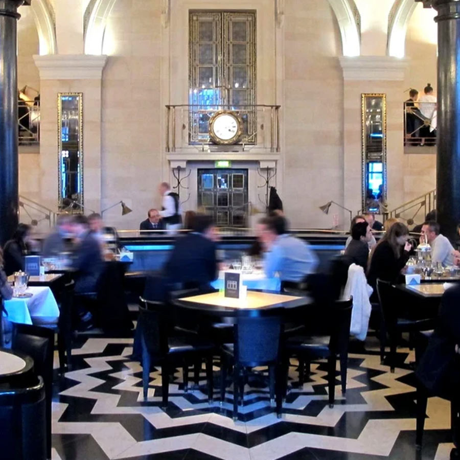 A bustling restaurant interior with patrons seated at tables, a large clock above the entrance, arched windows, mirrors, and a bold black-and-white geometric patterned floor.