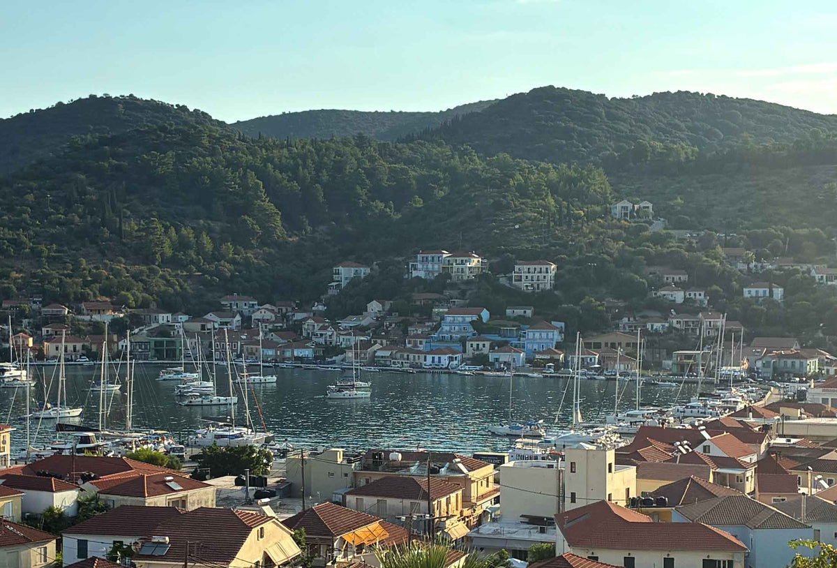Scenic coastal harbor in Ithaca, Geeece, with numerous white sailboats and yachts in calm blue water, surrounded by dense buildings with terracotta roofs and backed by steep, forested green hills under a pale blue sky.